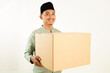 © Wayan - An Indonesian Muslim man with a happy expression and holding a large cardboard box isolated on a plain white background, wearing traditional Islamic clothing