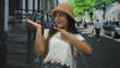 © Krakenimages.com - Young woman in straw hat points finger to palm and smiles on a city street while wearing white embroidered top and denim jeans; playful.