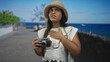 © Krakenimages.com - Young hispanic woman holding vintage camera, hand on chest gesture on a seaside street; concern travel curiosity.