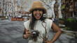 © Krakenimages.com - Young woman with straw hat holding vintage camera, hand on hip and smiling in a busy street market; travel joy adventure.