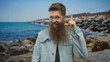 © Krakenimages.com - Man wearing denim jacket smiling and pointing finger skyward on a sunlit rocky beach shore by turquoise sea; optimism.