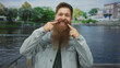 © Krakenimages.com - Man with long reddish beard wearing denim jacket points finger to mouth on street overlooking river canal guardrail; happiness positivity.