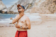 © qunica.com - A shirtless man wearing a cap and sunglasses applies sunscreen on a sunny beach. The scene shows red shorts and a tattooed arm, with rugged cliffs and calm waves in the background.