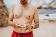 © qunica.com - A shirtless man on a sandy beach uses a sunscreen spray bottle, preparing for sun exposure. Calm seaside scene with rocks and waves in the background; lifestyle, outdoor, health, and summer vibe.