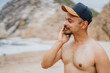 © qunica.com - A shirtless man wearing a cap on a sandy beach, applying sunscreen, with rocky cliffs in the background. A relaxed, sunny moment perfect for travel, lifestyle, or vacation themes.
