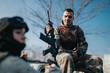 © qunica.com - Two stock-style sentences: A camouflaged soldier sits with an airsoft rifle in hand. The outdoor scene shows a teammate blurred in the background during a paintball-style game.