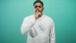 © Krakenimages.com - Hispanic man with finger to lips for silence in studio, beard and earring visible, white shirt against teal backdrop; calm introspection.