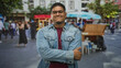 © Krakenimages.com - Hispanic man with glasses and beard standing with arms crossed, hand on sleeve, denim jacket, street market stalls and umbrella behind him; confidence casual.