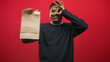 © Krakenimages.com - Hispanic man holding a brown paper bag while smiling and making an ok sign over his eye with his bare hand against a red seamless backdrop in studio; cheerful delivery promotion.