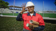 © Krakenimages.com - Man construction worker in hard hat and red safety jacket holding rolled blueprints and making ok sign while standing by stadium field in building; confidence planning safety.