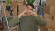 © Krakenimages.com - Smiling man making heart shape with hands in building among unpacked cardboard boxes and ladder with potted plant visible; love warmth.