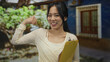 © Krakenimages.com - Young chinese woman outdoors holding clipboard while gesturing strength and invitation, surrounded by nature and traditional architecture, exuding confidence and friendliness.