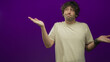 © Krakenimages.com - Hispanic man in beige t shirt with palms up shrugging against purple studio backdrop, visible wristbands, ring, piercing and tousled hair; uncertainty ambiguity.