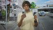 © Krakenimages.com - Man holding smartphone and creditcard in hand on street, comparing cards while frowning at the screen near parked cars and a building facade; indecision.