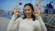 © Krakenimages.com - Woman smiling joyfully making ok gesture with both hands in a lively seaside port setting with boats visible in the background on a sunny day.