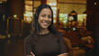 © Krakenimages.com - Hispanic woman with arms crossed smiles confidently indoors at a stylish hotel lounge, creating a warm and welcoming atmosphere.