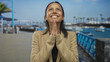 © Krakenimages.com - Hispanic woman smiling joyfully outdoors at a seaside port with boats in sight, clapping hands against a coastal backdrop creating a sense of happiness and tranquility.