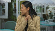 © Krakenimages.com - Young hispanic woman appears concerned on a street terrace, outdoors, wearing a jacket, showcasing urban lifestyle and emotions in a public setting with tables and chairs around.