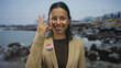 © Krakenimages.com - Hispanic woman smiling outdoors by the seaside, wearing an 'i voted' badge with usa flag, gesturing with her fingers, capturing a joyful and patriotic moment at the beach.