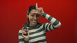 © Krakenimages.com - Young hispanic woman with a remote control smiling and making a loser gesture on a vibrant red wall background in an isolated and playful setting.