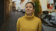 © Krakenimages.com - Woman smiling in a cozy yellow sweater stands confidently on an urban street, embodying tranquility and warmth in a rustic alley setting.