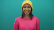 © Krakenimages.com - Hispanic woman in a pink sweater and yellow hat making a secret gesture against a vibrant green background, exuding youthfulness and playful curiosity.