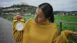 © Krakenimages.com - Woman in yellow sweater holds alarm clock in park expressing confusion with green field and cloudy sky in background.