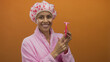 © Krakenimages.com - Hispanic woman in a pink bathrobe and patterned shower cap holds a razor against an orange background, smiling and appearing content with her personal care routine.
