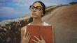 © Krakenimages.com - Young hispanic woman reading a book outdoors with a serene expression, wearing glasses and a sleeveless blouse against a scenic background of a dirt path under a blue sky.