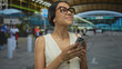 © Krakenimages.com - Woman smiling while using smartphone at busy airport terminal outdoors with modern architecture around her.