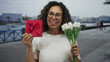 © Krakenimages.com - Woman smiling by seaside promenade holding flowers and a gift box in an outdoor setting, capturing a joyful moment by the beach.