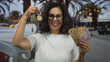 © Krakenimages.com - Middle-aged hispanic woman holds a house key and mexican pesos outdoors, symbolizing real estate and financial success in an urban setting.