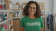 © Krakenimages.com - Woman smiling in charity room wearing volunteer shirt illustrating hispanic female volunteer in indoor community setting with organized shelves showcasing charity supplies.