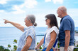 © luciano - Happy senior couple with a female friend looking at the horizon while enjoying a scenic ocean view from a seaside promenade. Three older adults standing together on a sunny day expressing friendship