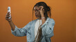 © Krakenimages.com - Woman holding smartphone and adjusting glasses for selfie in studio orange background; self expression confidence.