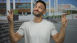© Krakenimages.com - Hispanic young man outdoors in an urban street making air quote gestures, expressing sarcastic disbelief in a casual white t-shirt against a blurred city background.