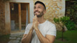 © Krakenimages.com - Young man in white shirt smiling with hands clasped in prayer in front of a rustic mediterranean house with stone walls and greenery.