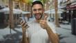 © Krakenimages.com - Hispanic man in white shirt gestures thumbs up while using smartphone on urban street with sunny ambiance showcasing modern tech interactions.