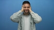 © Krakenimages.com - Young man covering ears in frustration against a solid blue background, expressing discomfort and stress, dressed in casual attire.