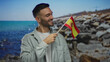 © Krakenimages.com - Young man holding spanish flag smiling at beach with blue sea and rocky shore in background embodying hispanic pride and outdoor enjoyment.