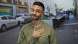 © Krakenimages.com - Young hispanic man standing on an urban street with cars and graffiti in the background, placing hand on chest in a friendly gesture.