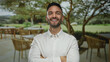 © Krakenimages.com - Young man with crossed arms and a bright smile stands on a terrace of an outdoor restaurant, surrounded by tables and lush landscape, embodying confidence and leisure.