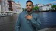 © Krakenimages.com - Man with hand on chest in denim shirt standing on street beside amsterdam canal and water with historic buildings along the quay, speaking with open mouth; sincerity trust.
