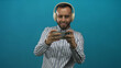 © Krakenimages.com - Young hispanic man holding smartphone and smiling, wearing headphones and striped shirt in teal studio for casual content creation; fun relaxation.