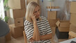 © Krakenimages.com - Woman sitting at table with hand on cheek and folded arm looks upward amid stacked cardboard moving boxes and houseplant in building; moving stress.
