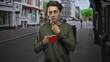 © Krakenimages.com - Handsome young man holding red mug, hand to chest on cobblestone urban street with blurred shops; contemplation solitude.