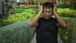 © Krakenimages.com - Chinese man in black shirt grimaces and holds head with both hands by red building wall; stress frustration.