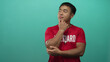© Krakenimages.com - Young chinese lifeguard man in red shirt with lifeguard print and whistle holds chin in studio with green wall; contemplation.