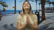 © Krakenimages.com - Woman praying with joyful expression on a sunny seaside promenade, showcasing a peaceful outdoor scene with palm trees and a clear blue sea in the background.