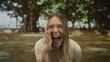 © Krakenimages.com - Young blonde woman expressing excitement and shouting in a sunny outdoor park setting with vibrant trees in the background.